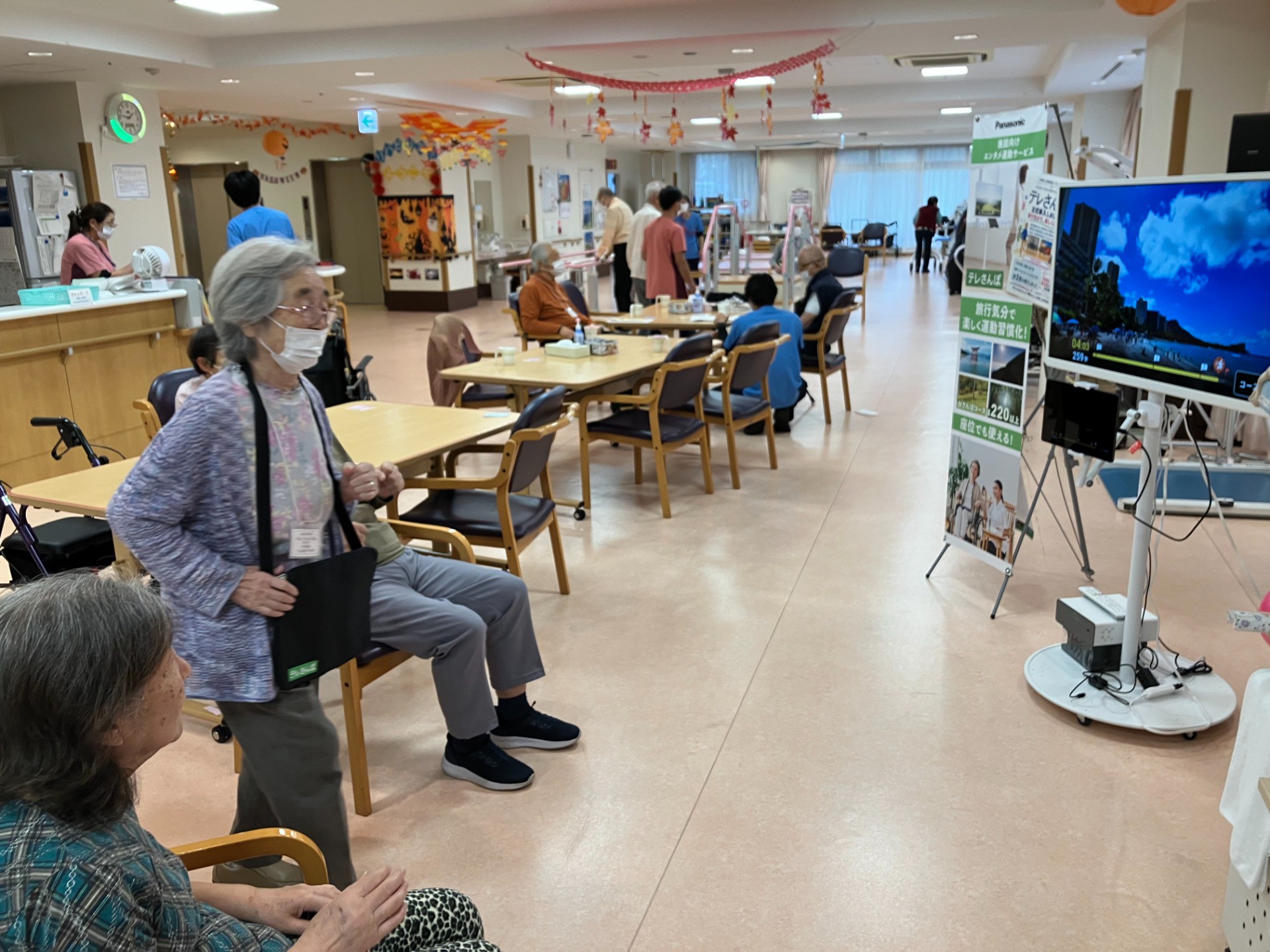 イルアカーサでパナソニック株式会社の「テレさんぽ」を使い、立位と座位でワイキキビーチの映像を見ながら運動する複数の利用者様irua-casa-panasonic-telesampo-waikiki-standing-seated.